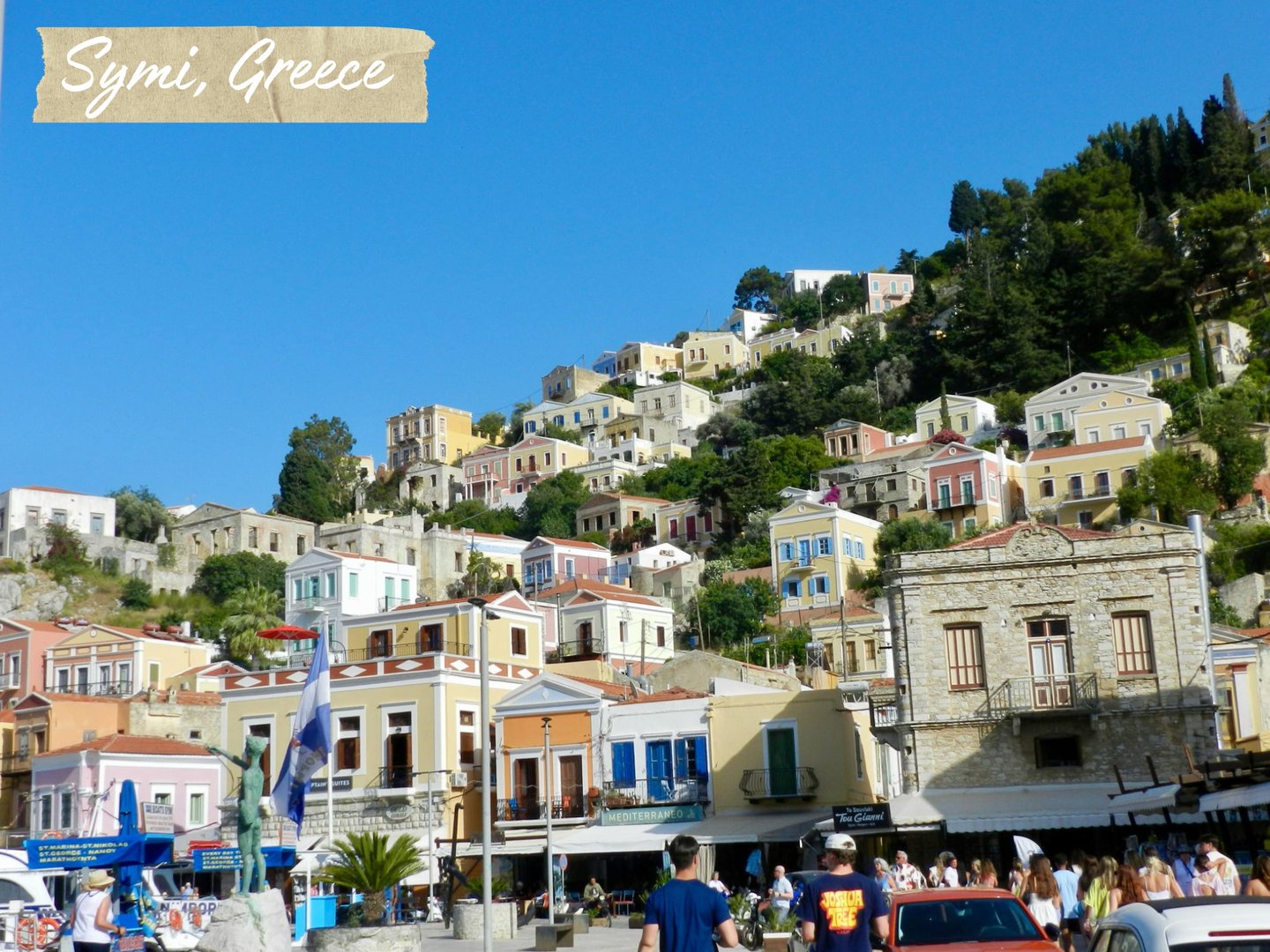 Colorful neoclassical houses on a hillside overlooking the harbor in Symi, Greece under a blue sky.