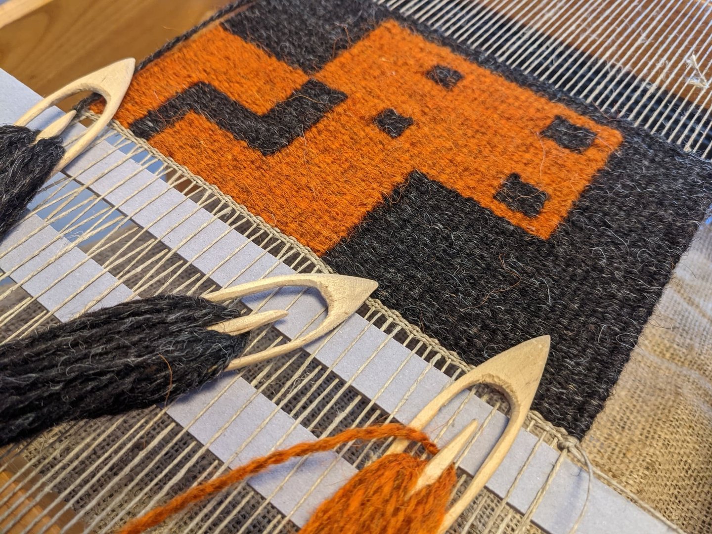 A weaving loom with shuttles and orange, yellow, and black wool threads on a kilim rug with a pixel-