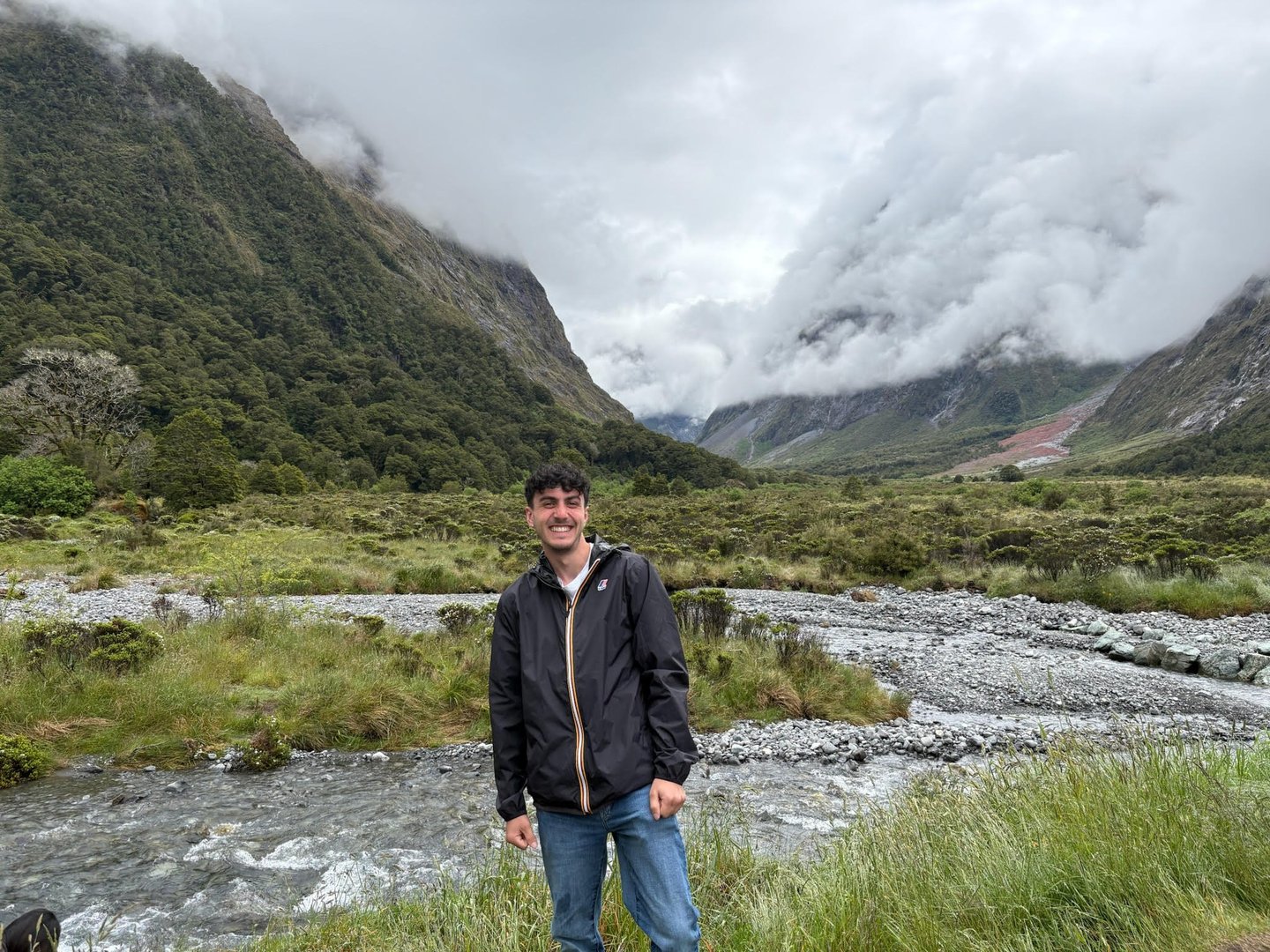 Outdoor portrait of Luca smiling in a mountain valley with a river and cloudy peaks in the backgrou