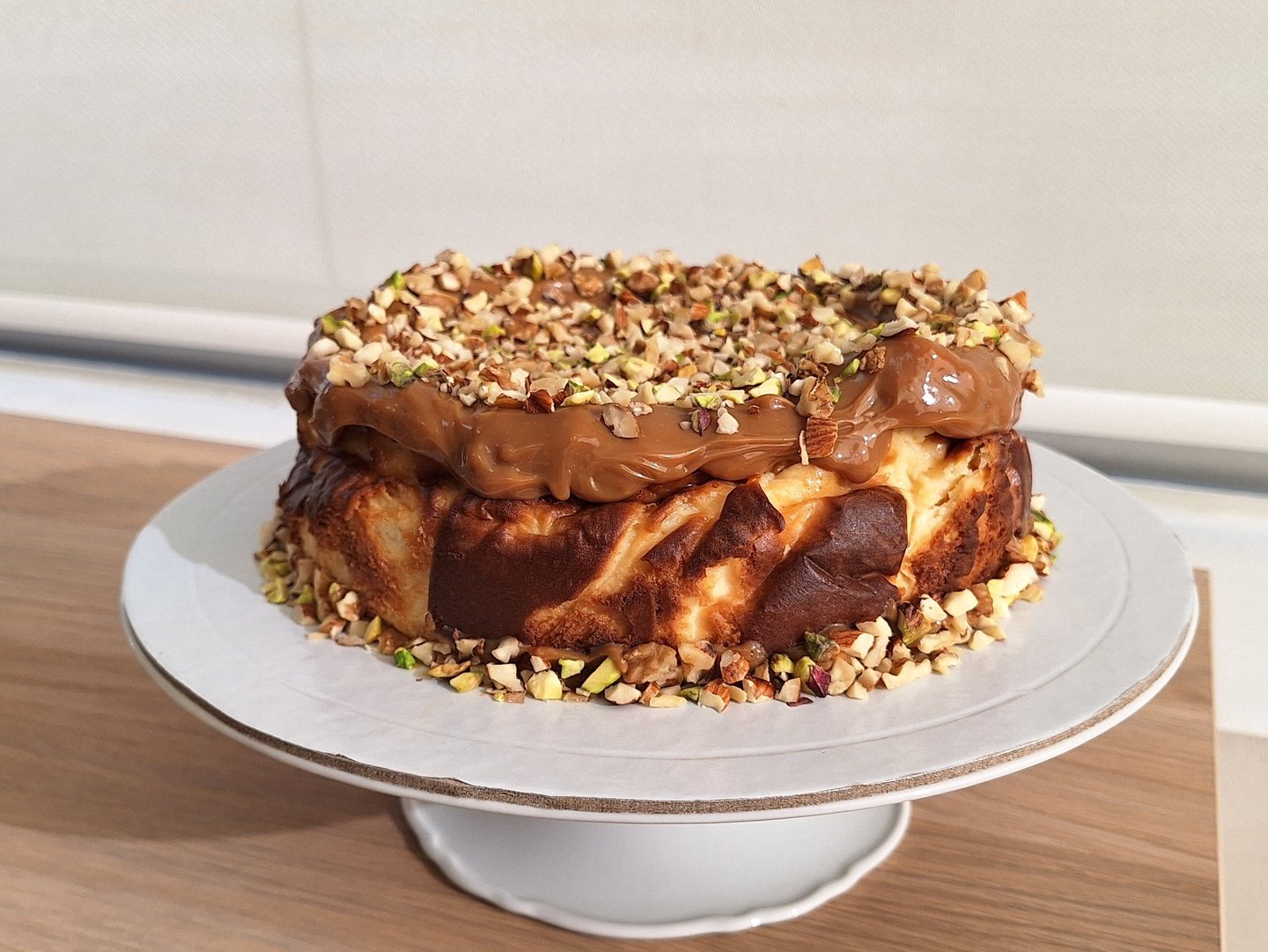 San Sebastian burnt cheesecake topped with creamy caramel and crushed pistachio nuts on a white cake stand.