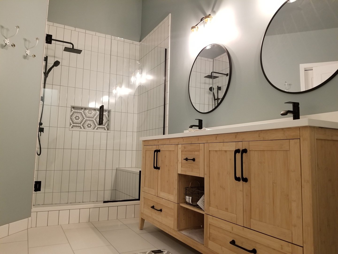 Modern bathroom featuring a light wood vanity, round black mirrors, and a walk-in glass shower with white tile.