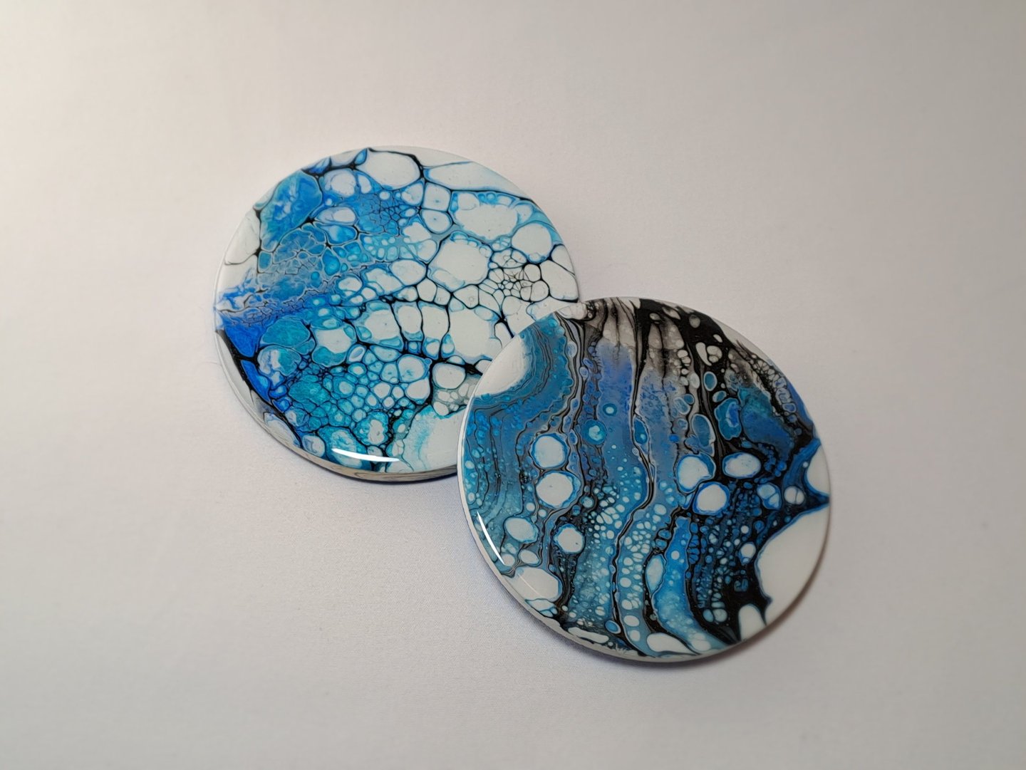 Two round acrylic pour coasters with blue, white, and black abstract cell patterns.
