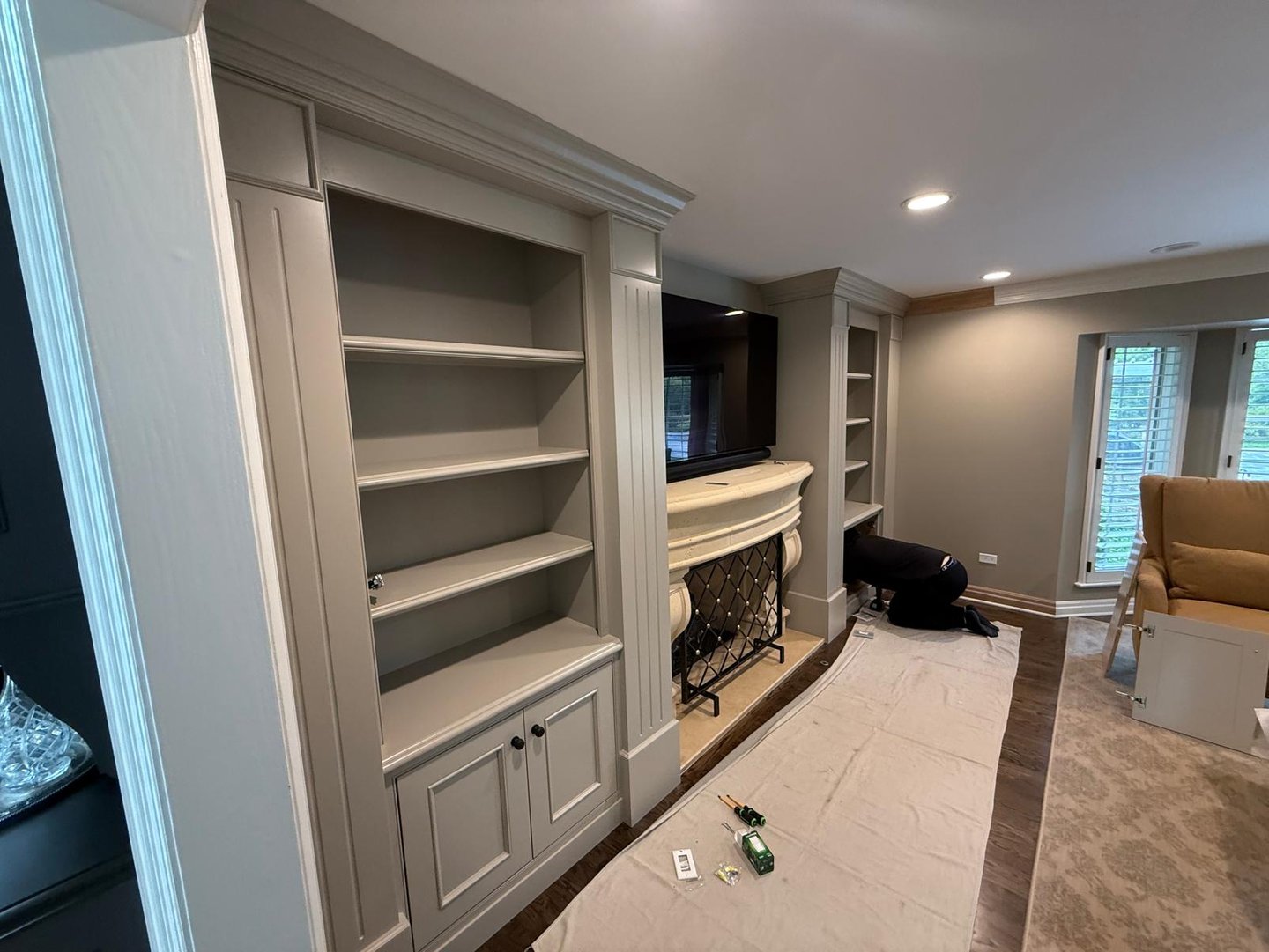A carpenter installs custom gray built-in bookshelves and cabinetry flanking a living room fireplace.