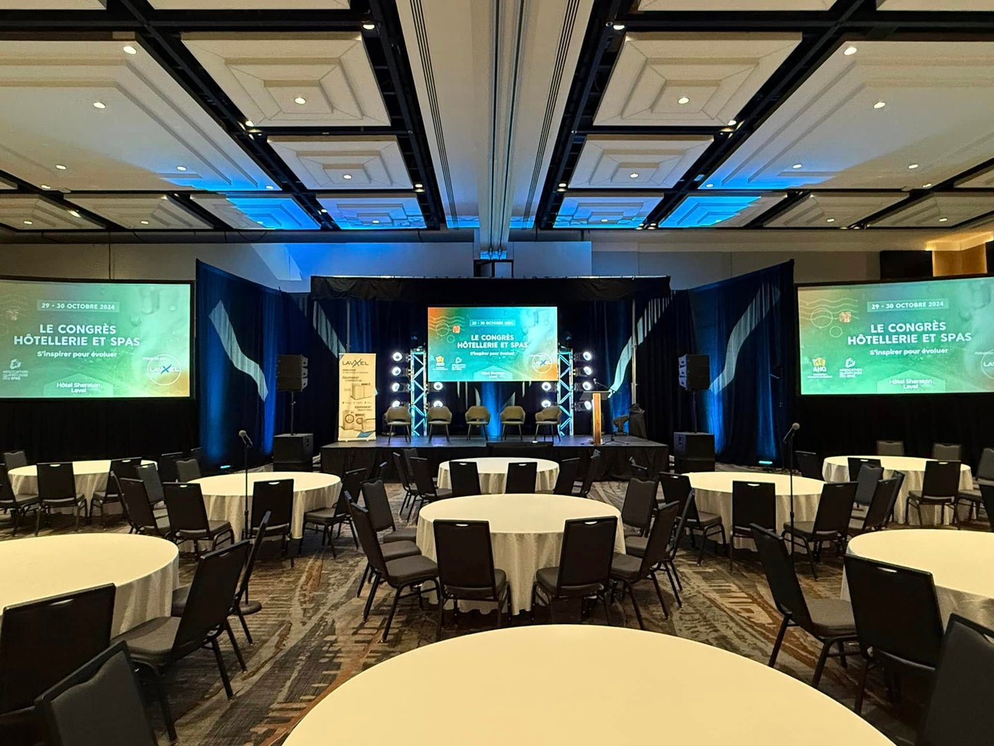 Conference room setup for the "Congrès Hôtellerie et Spas" at the Sheraton Laval, featuring a stage with large screens