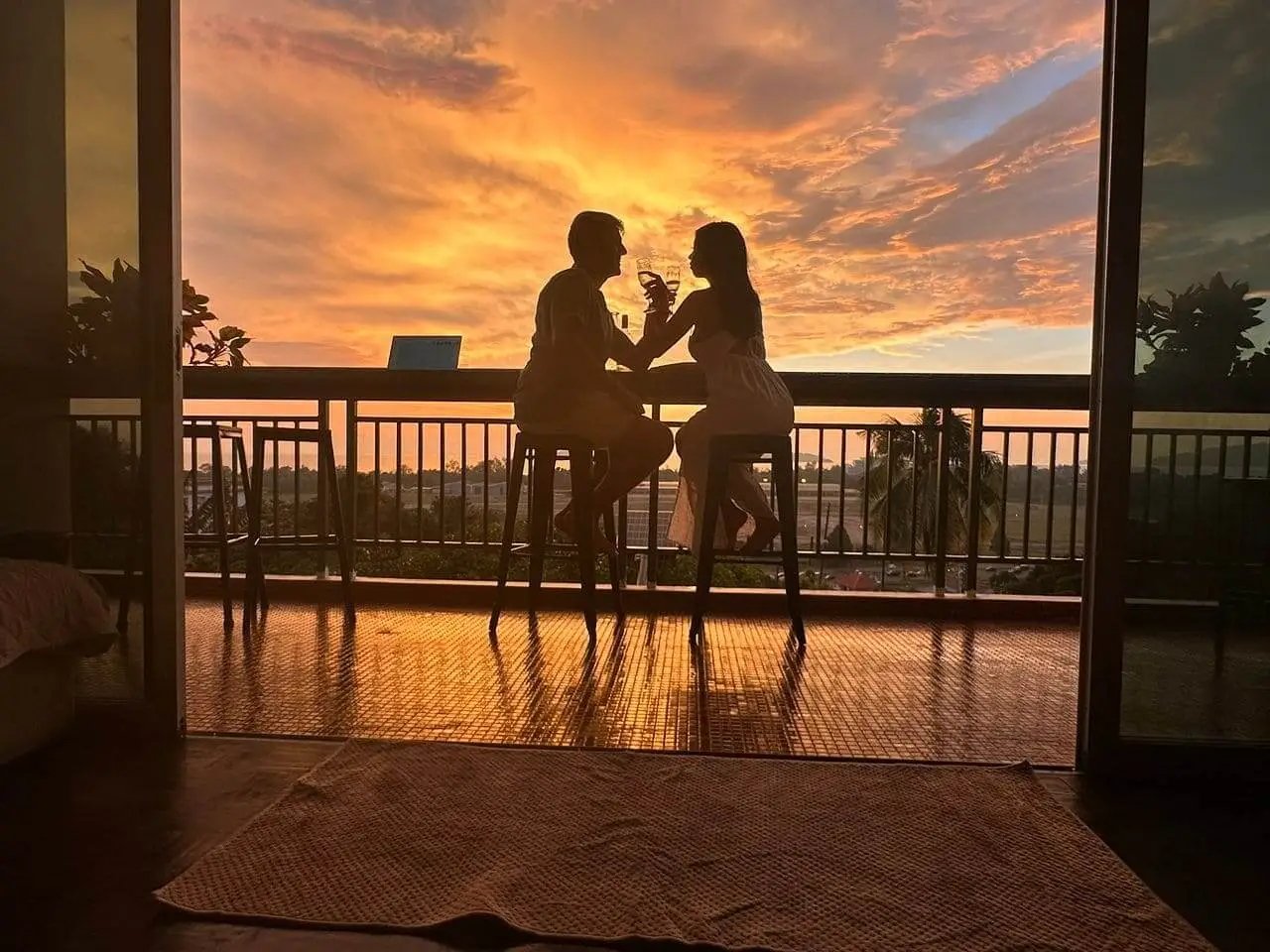 Silhouette of a couple enjoying drinks on a balcony at sunset with a vibrant orange sky.