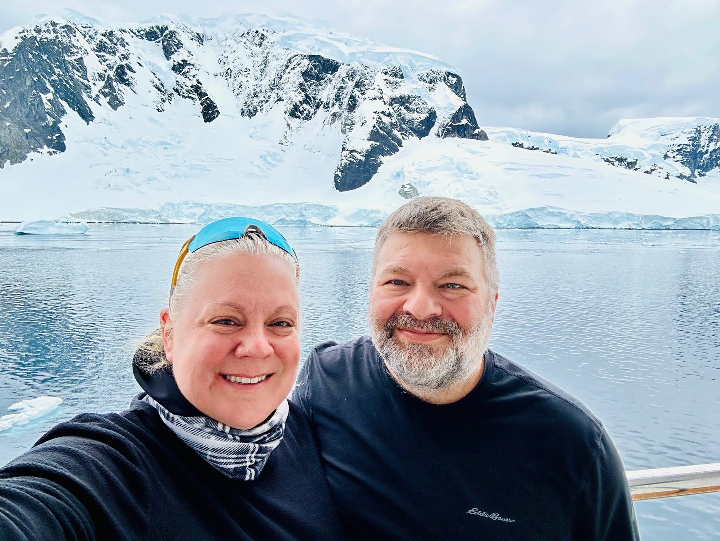 Smiling couple taking a selfie on an Antarctica cruise with snowy mountains and glaciers in the background.