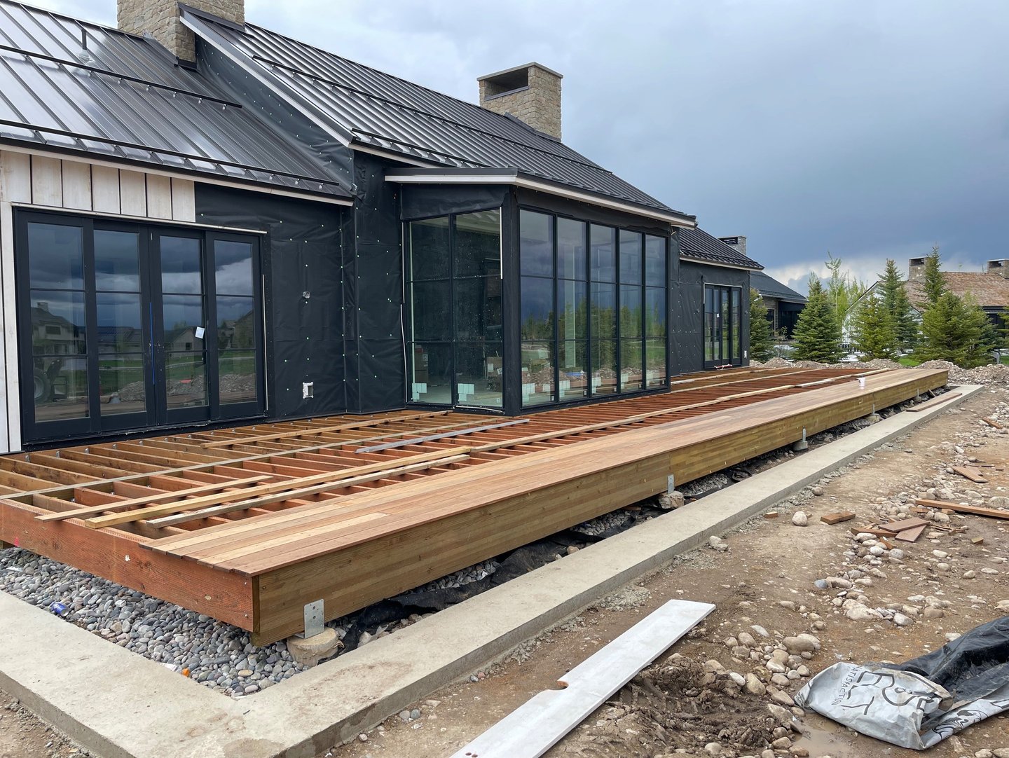 A modern luxury home under construction featuring a new wooden deck frame and floor-to-ceiling glass windows.