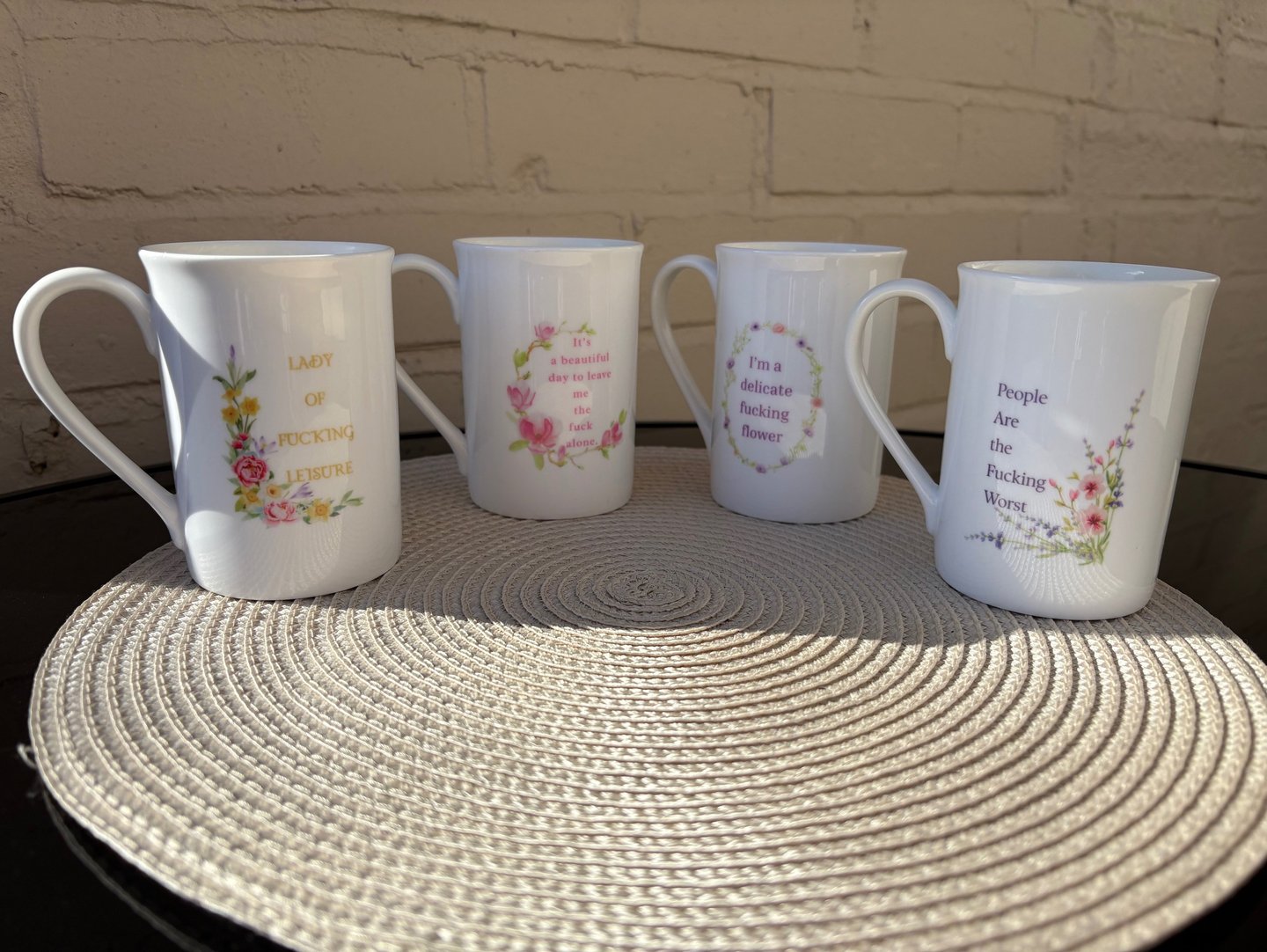 Four white bone china mugs with floral designs and funny, profane quotes on a woven placemat.