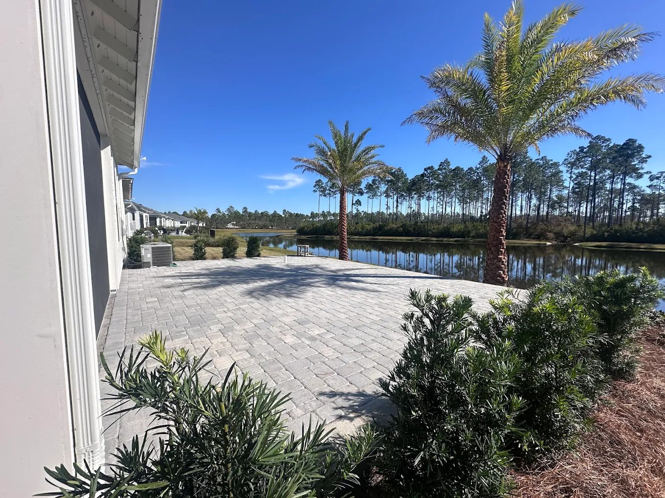 A large stone paver patio overlooking a scenic lake with palm trees and a clear blue sky.