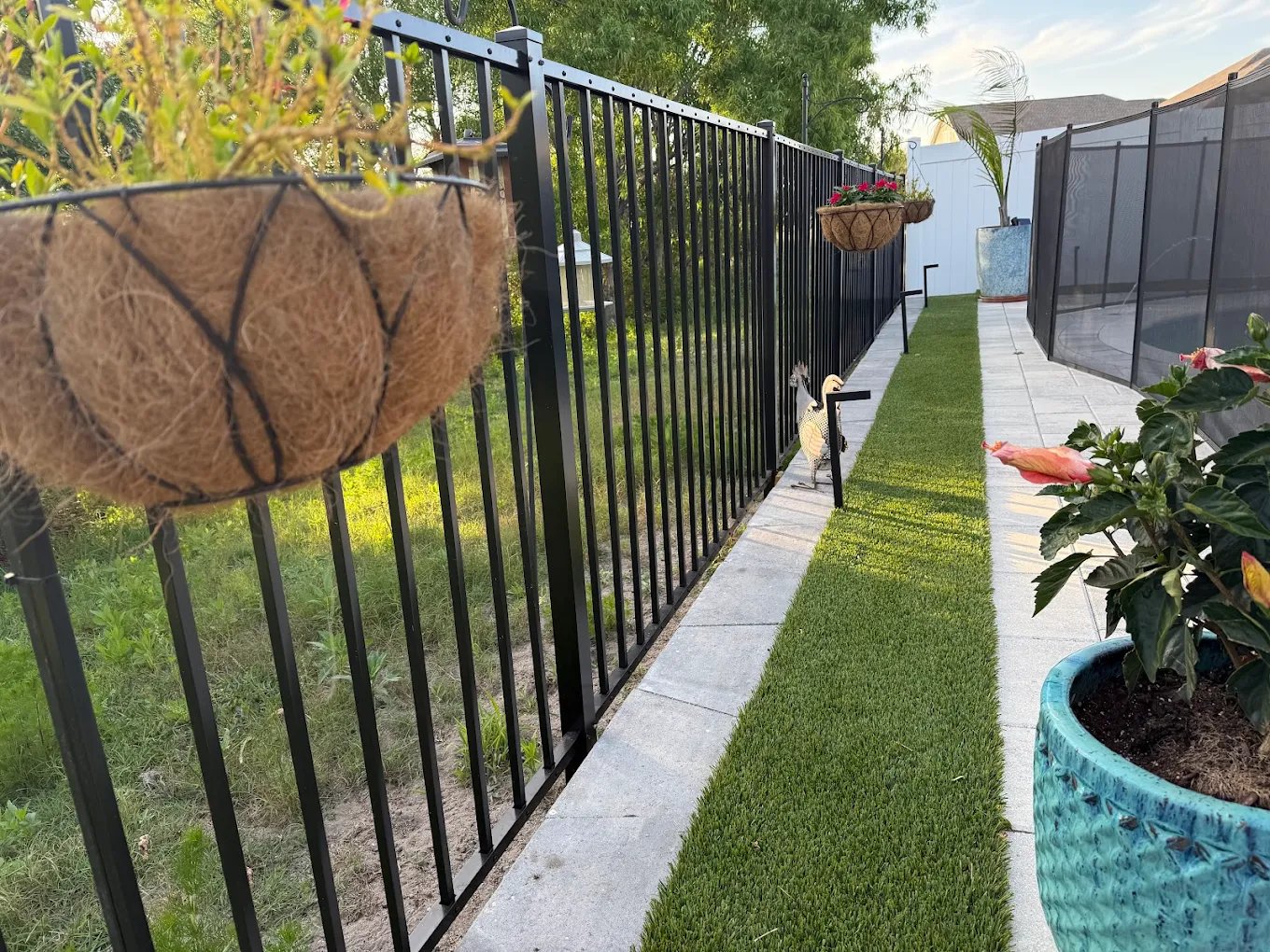 Black metal pool fence with hanging flower baskets and artificial grass turf walkway.