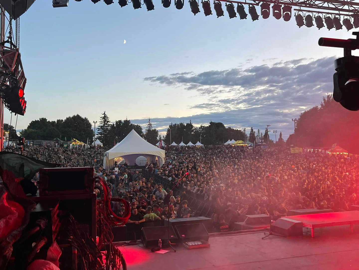 Crowd of fans at an outdoor music festival concert during a sunset with stage lighting.