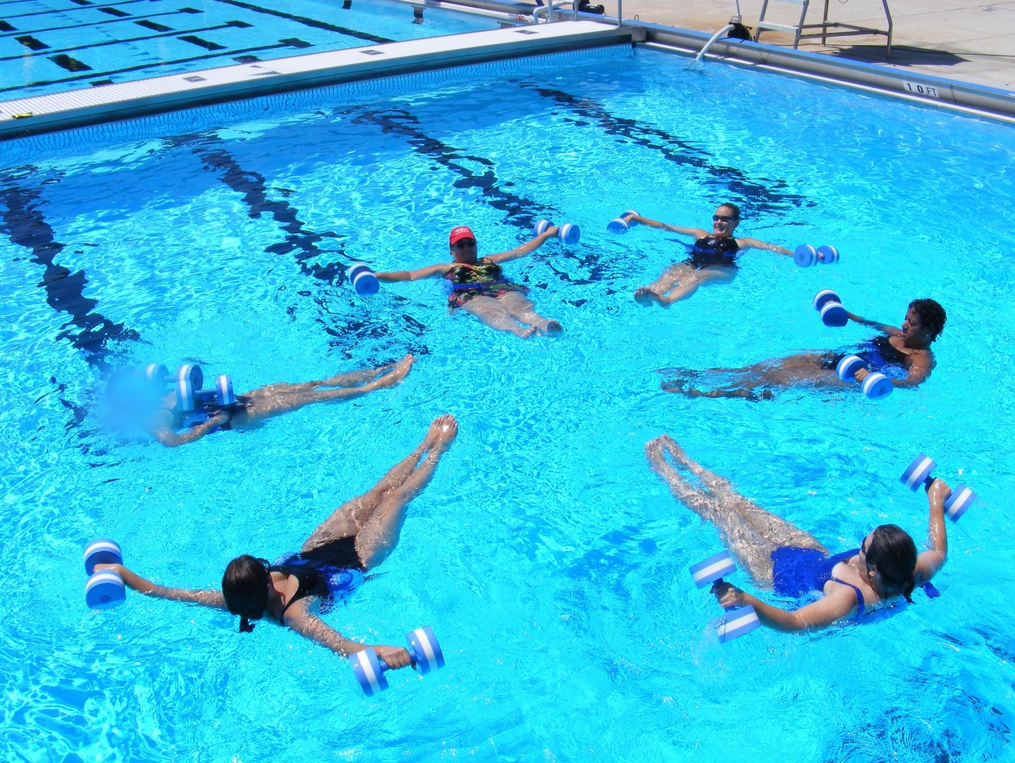 A group of women floating in an outdoor swimming pool while performing a water aerobics exercise with blue foam dumbbells.