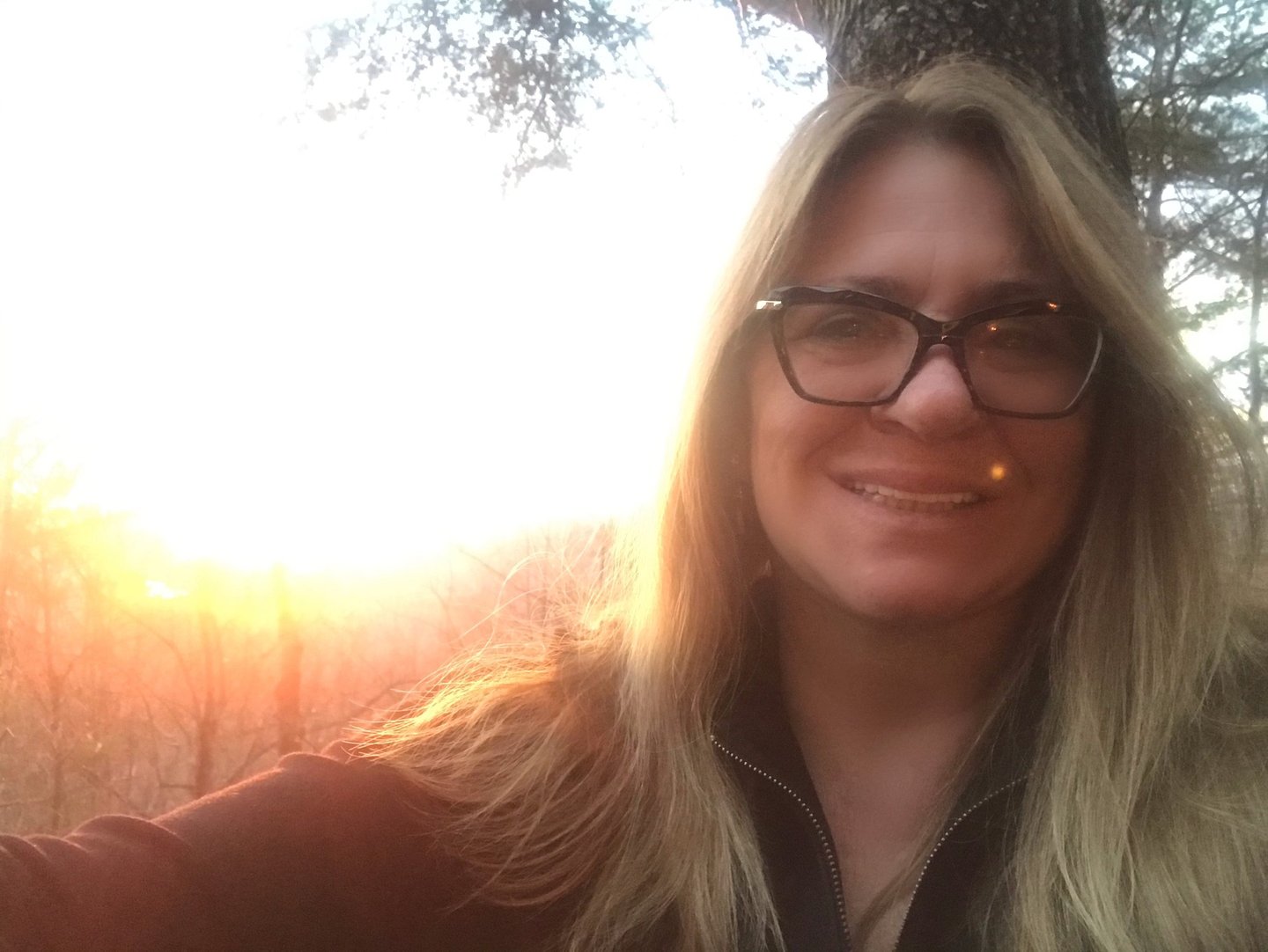 Smiling blonde woman with glasses taking a golden hour selfie during a scenic forest sunset.