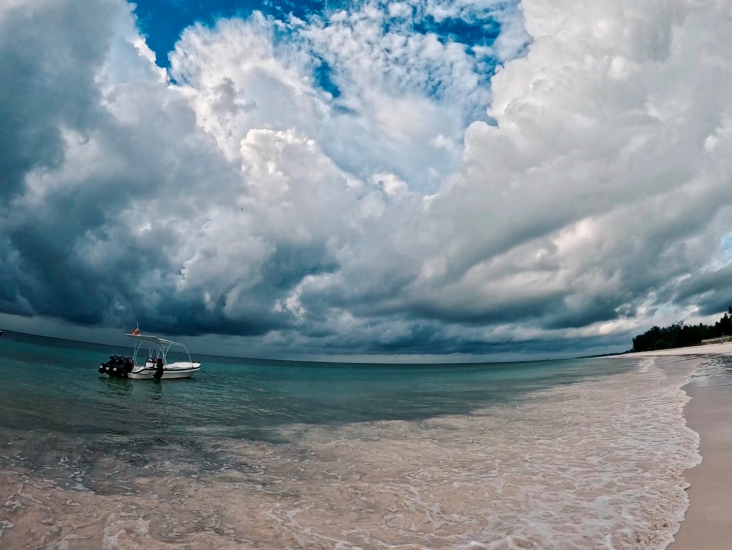 A small motorboat anchored near a tropical beach under dramatic storm clouds and a blue sky.