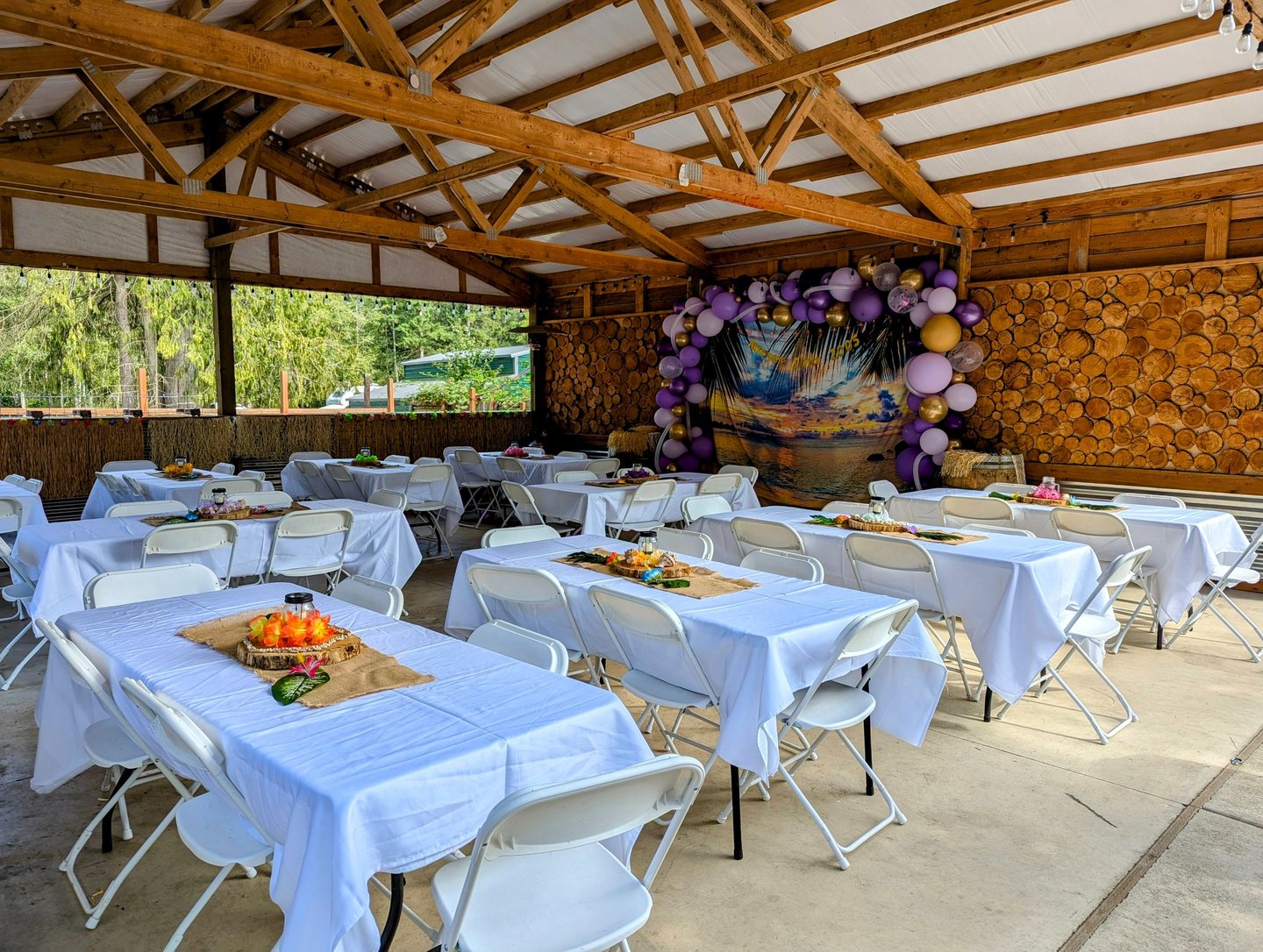 Rustic outdoor event venue featuring decorated banquet tables under a wooden pavilion with a tropical balloon arch.