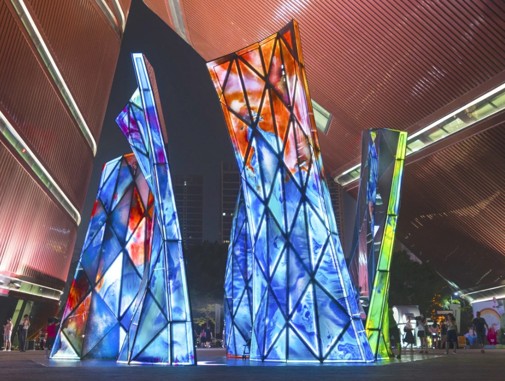 Blooming Sculpture lit up at night with its multi-colored glass panels and people walking around it