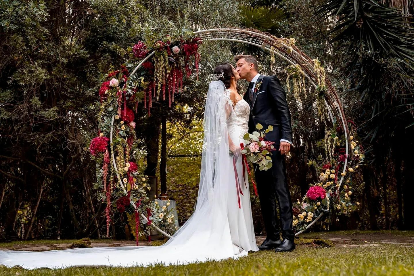 Bride and groom sharing kiss in front of circular wedding arch decorated with red and white flowers during outdoor ceremony.