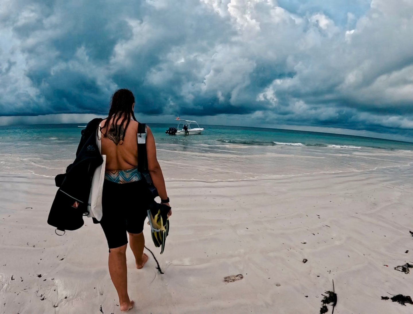 A scuba diver walks across Diani Beach's white sand beach toward a boat under dramatic storm clouds.