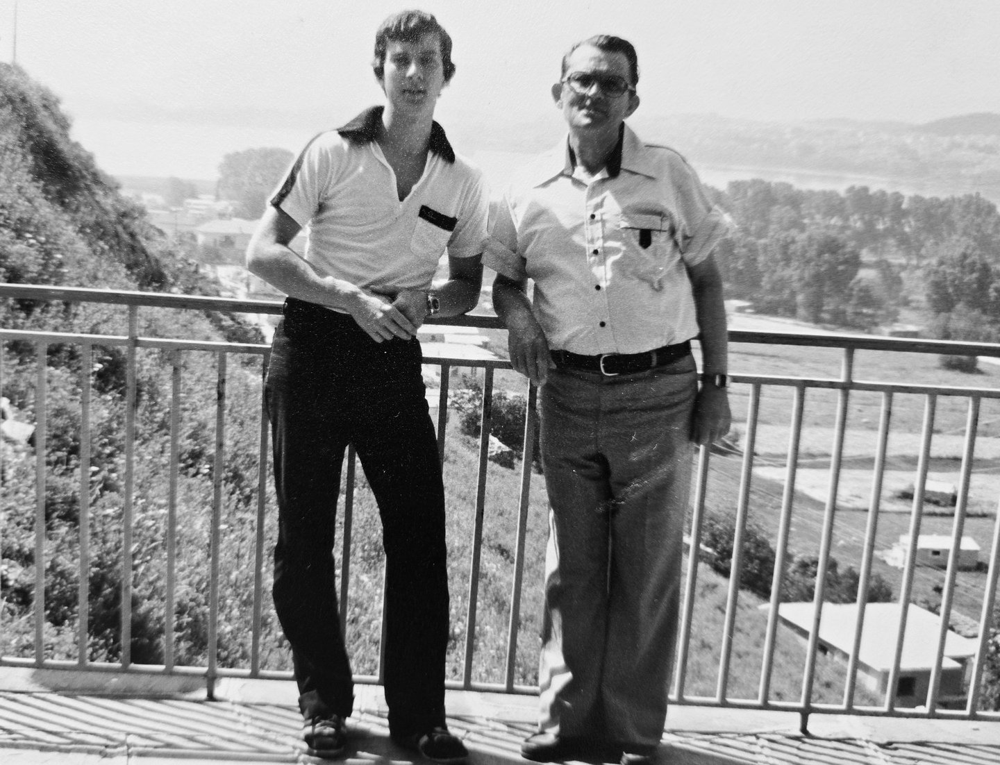 two men standing on a balcony with a view of a Greek city