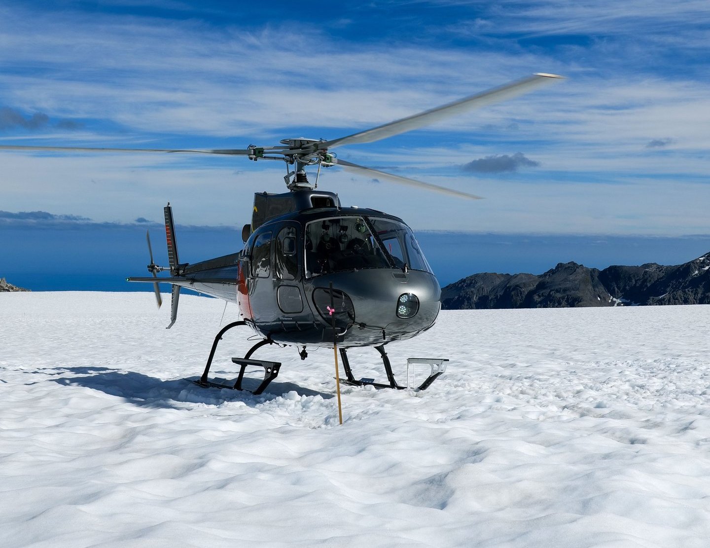 A grey scenic tour helicopter landed on a snowy mountain glacier under a blue sky.