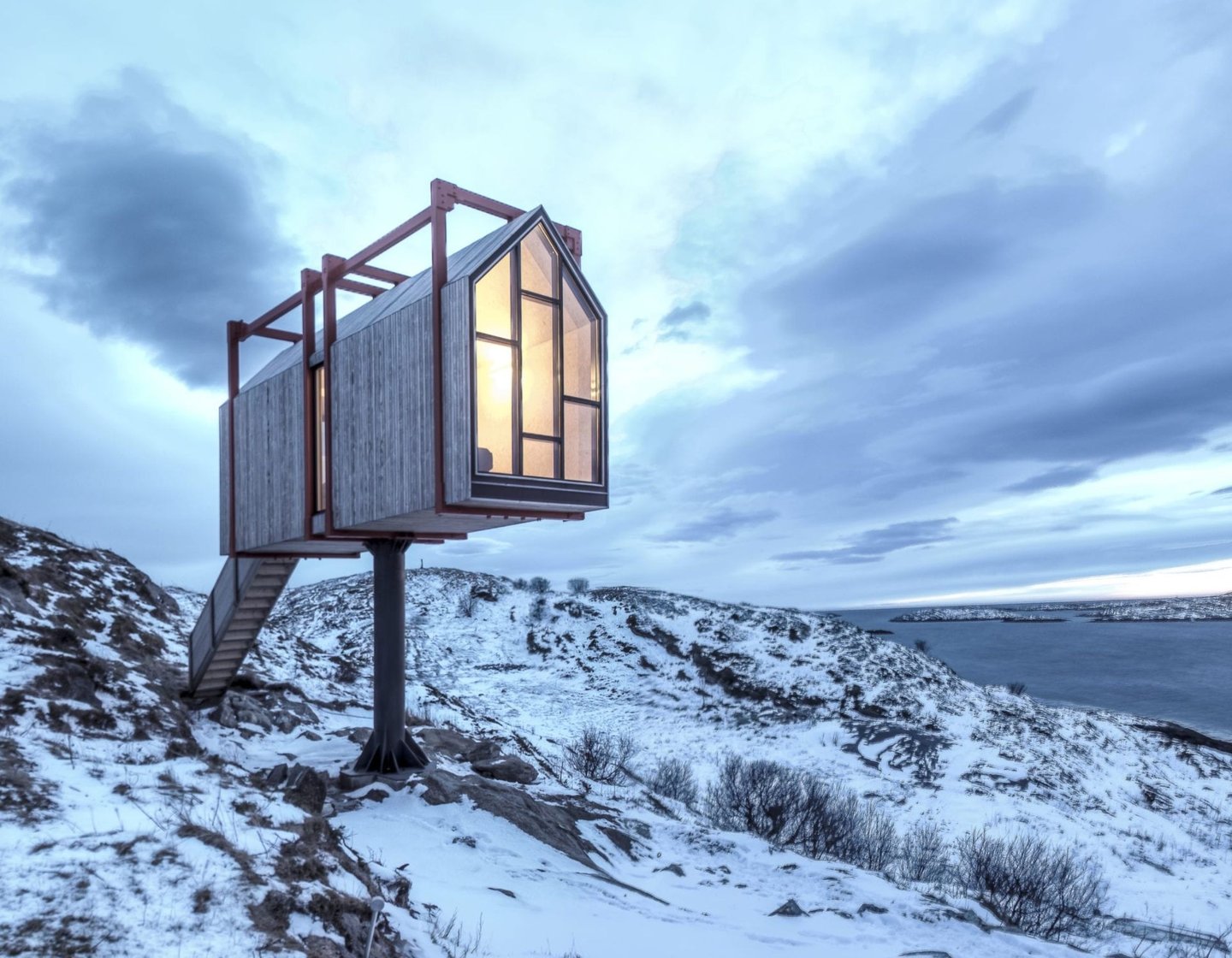 A modern elevated wooden cabin perched on a snowy hillside overlooking a lake under a cloudy sky.