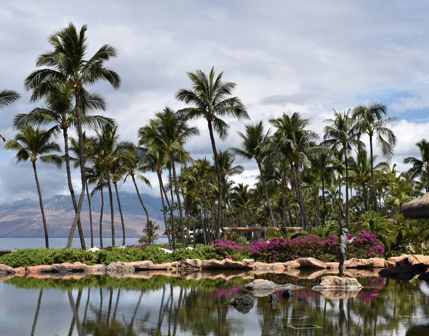 Tropical Hawaiian landscape featuring tall palm trees reflected in a calm resort pond with distant mountain views.