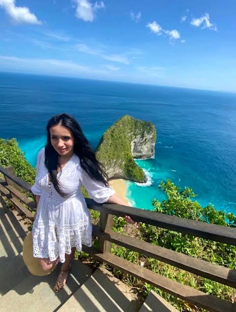 A young woman in a white lace dress poses at Kelingking Beach viewpoint in Nusa Penida, Bali.