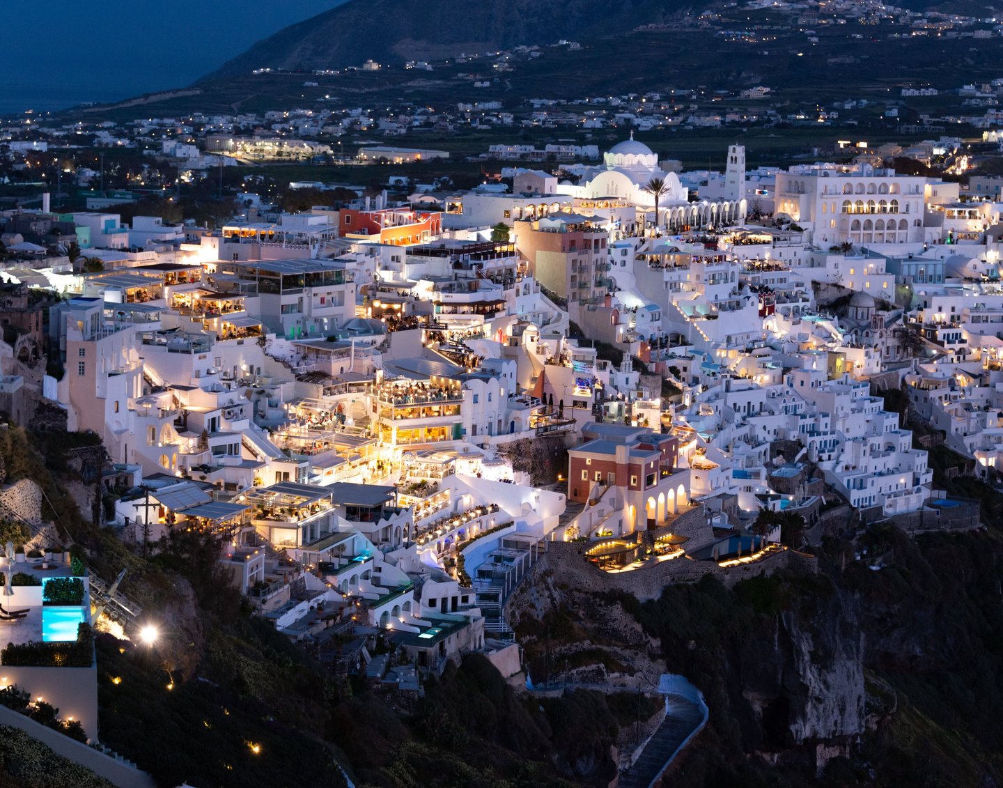 Night view of illuminated white buildings and cliffs in Fira, Santorini, Greece overlooking the Aegean Sea.
