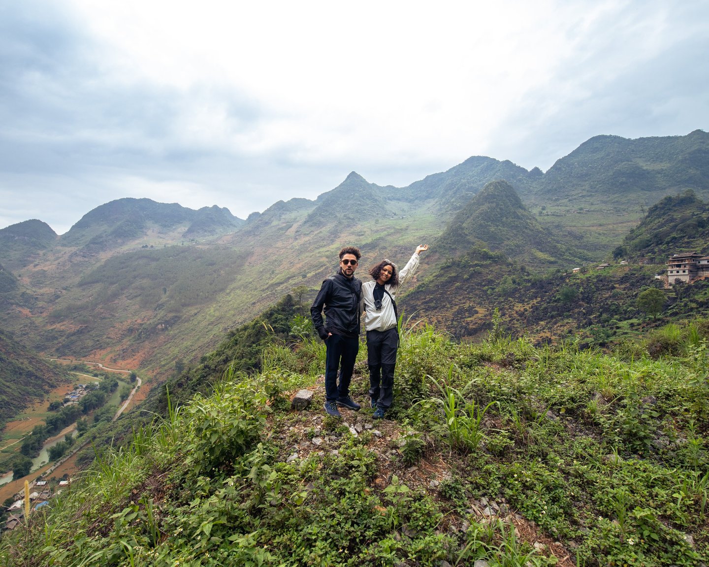 A couple standing on a grassy mountain edge posing for the camera. in the background are the beautiful Ha Giang mountains. 