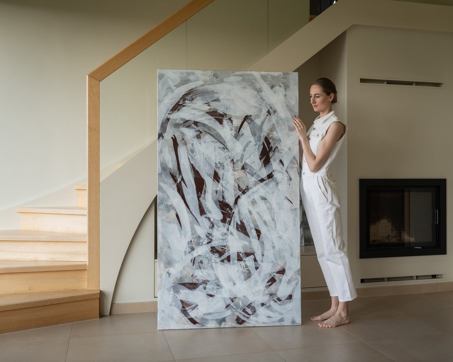 A female artist in white attire presents a large abstract painting in a modern home interior.