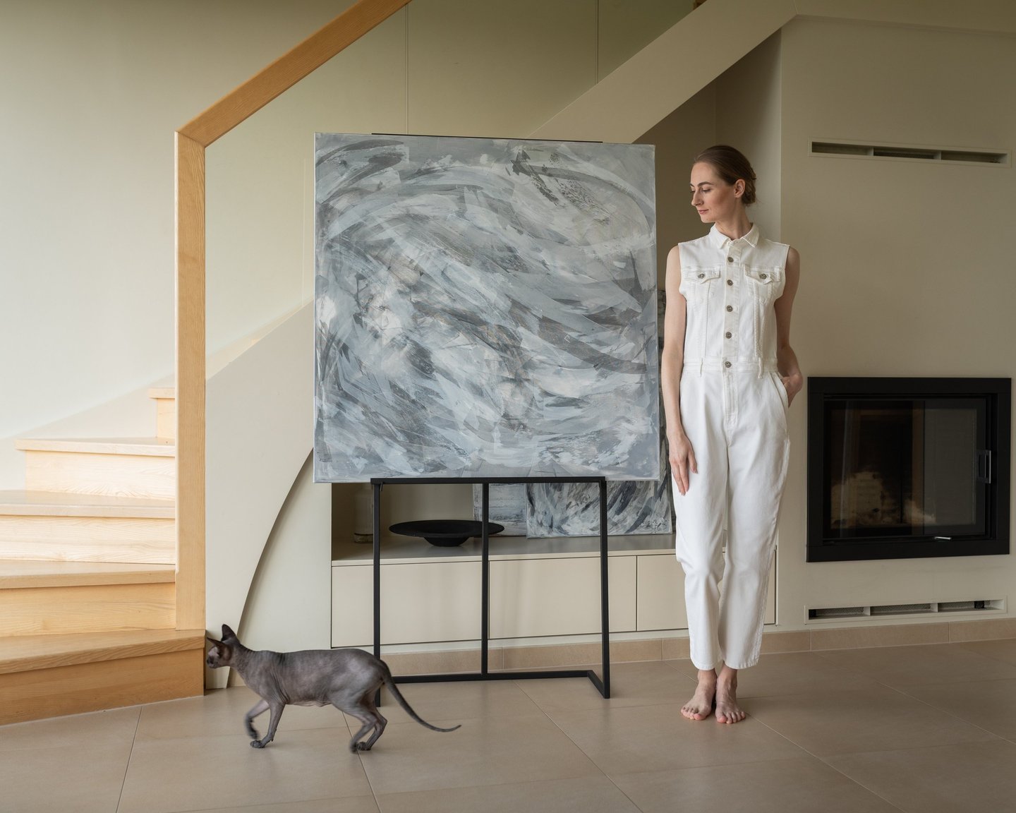 A woman in a white jumpsuit stands by abstract canvas art and a Sphynx cat in a modern home.