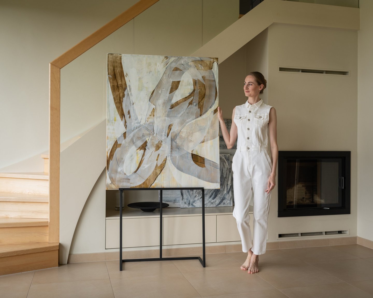 Artist in a white jumpsuit posing with a large abstract canvas painting in a modern home.