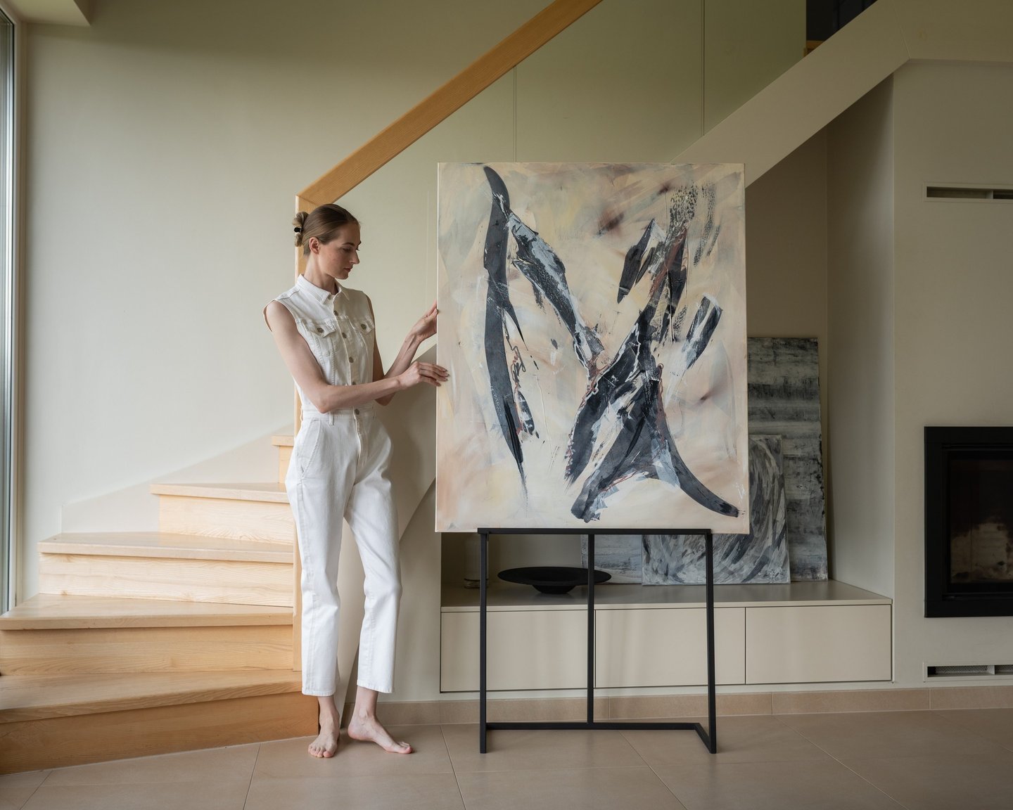 A female artist in a white jumpsuit poses beside a large abstract canvas painting on an easel in a modern minimalist home.