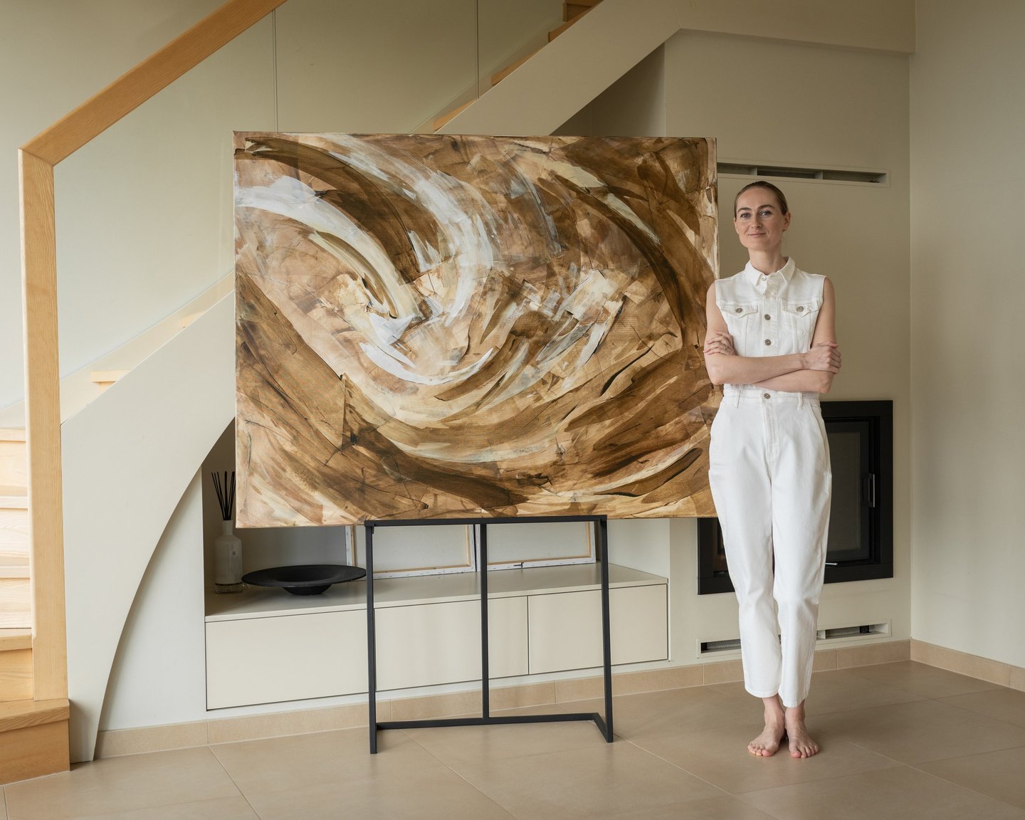 A female artist in a white jumpsuit standing next to her large abstract brown and beige painting on an easel.