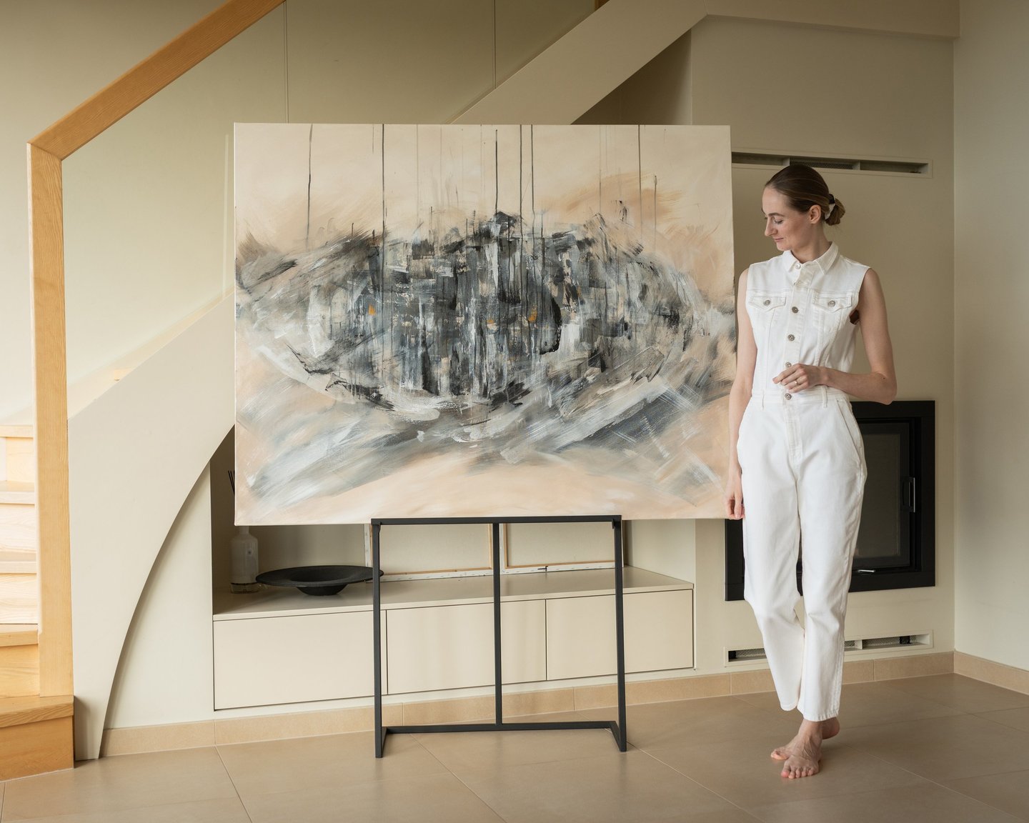 A woman in a white jumpsuit stands next to a large abstract expressionist painting in a modern home.