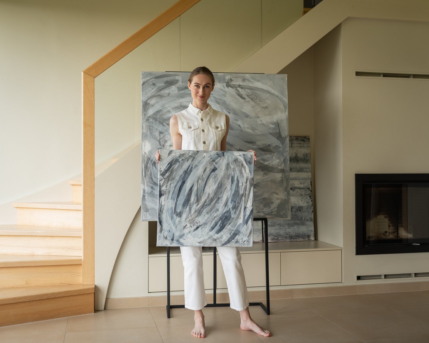 A smiling artist in a modern home holding a grey and white abstract canvas painting near a staircase.