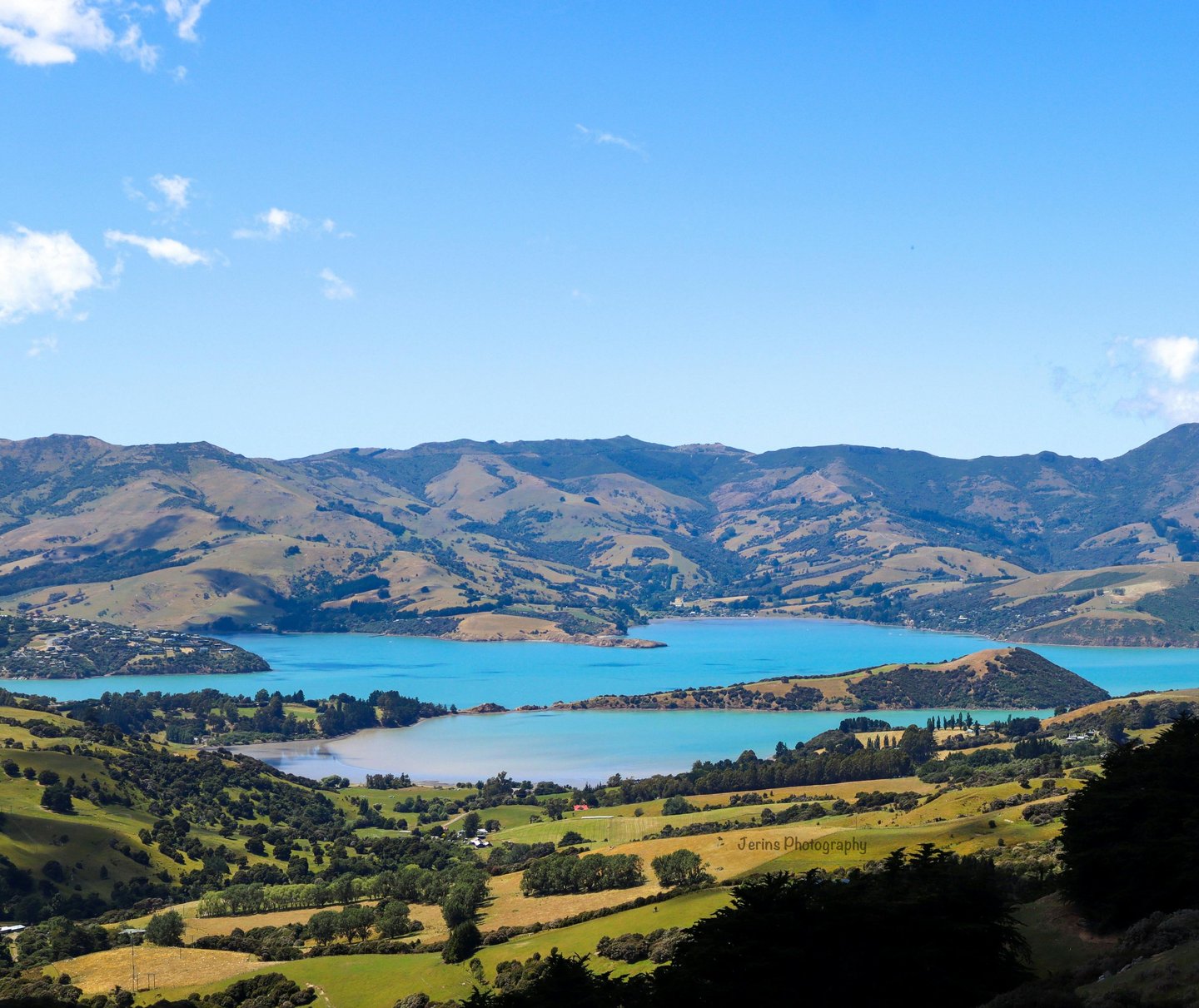 Panoramic view of Akaroa Harbour in New Zealand with turquoise water, rolling green hills, and mountains.