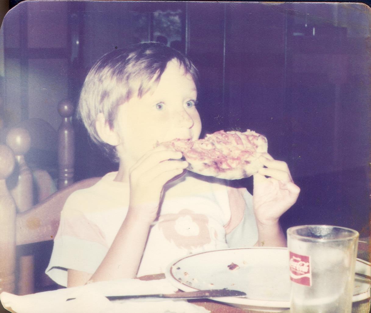 Vintage photo of a young boy eating a slice of pizza at a dinner table.