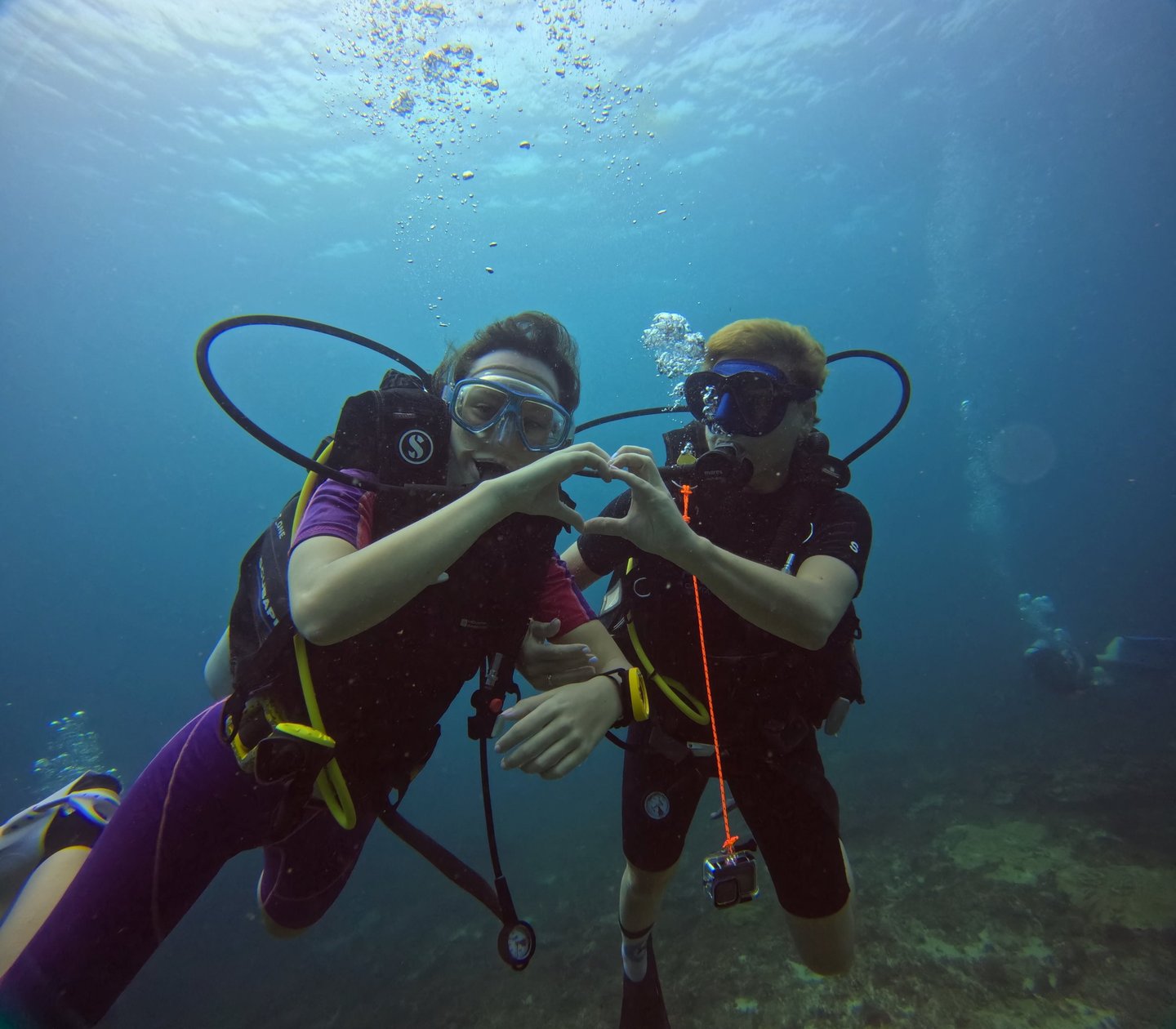 Two scuba divers making a heart shape with their hands while underwater diving in clear blue ocean water.
