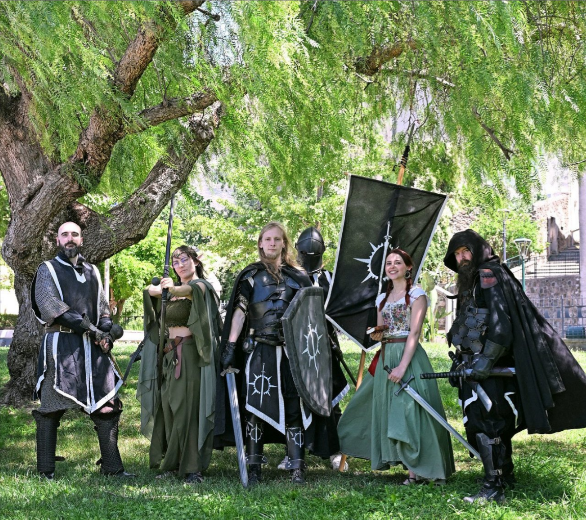 A group of medieval fantasy cosplayers in armor and robes holding swords and a banner under a large tree.