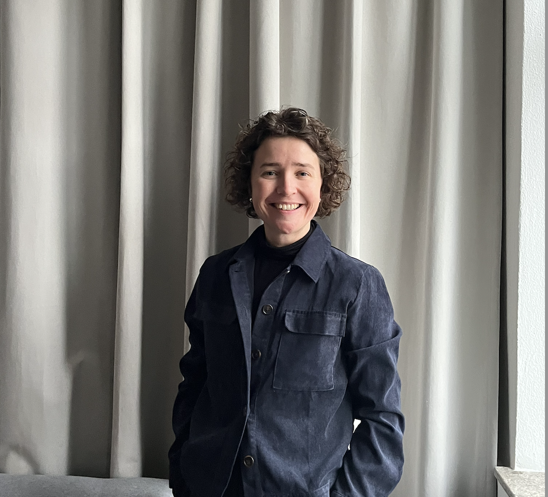 Smiling woman with curly hair wearing a blue jacket standing against light grey curtains.