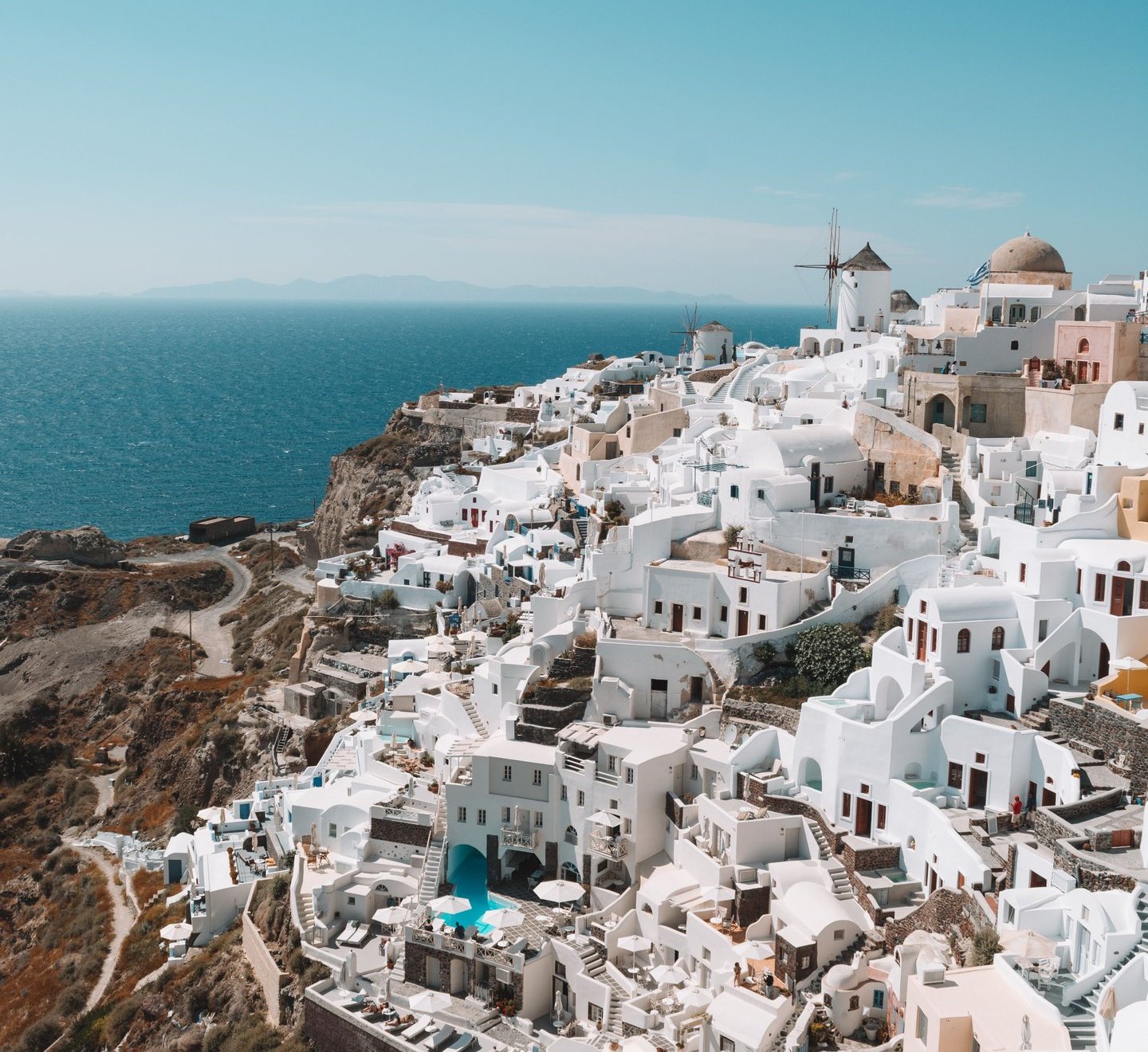 Panoramic view of white houses and a windmill on the cliffs of Oia, Santorini, overlooking the blue Aegean Sea.