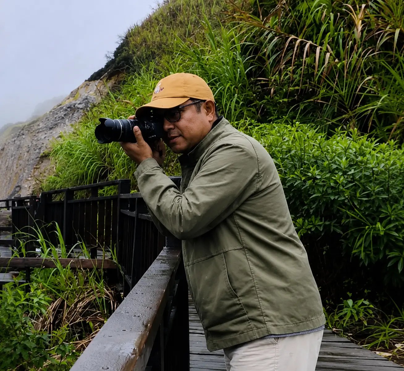 Photographer in a green jacket taking a picture with a DSLR camera on a wooden walkway in the mountains.