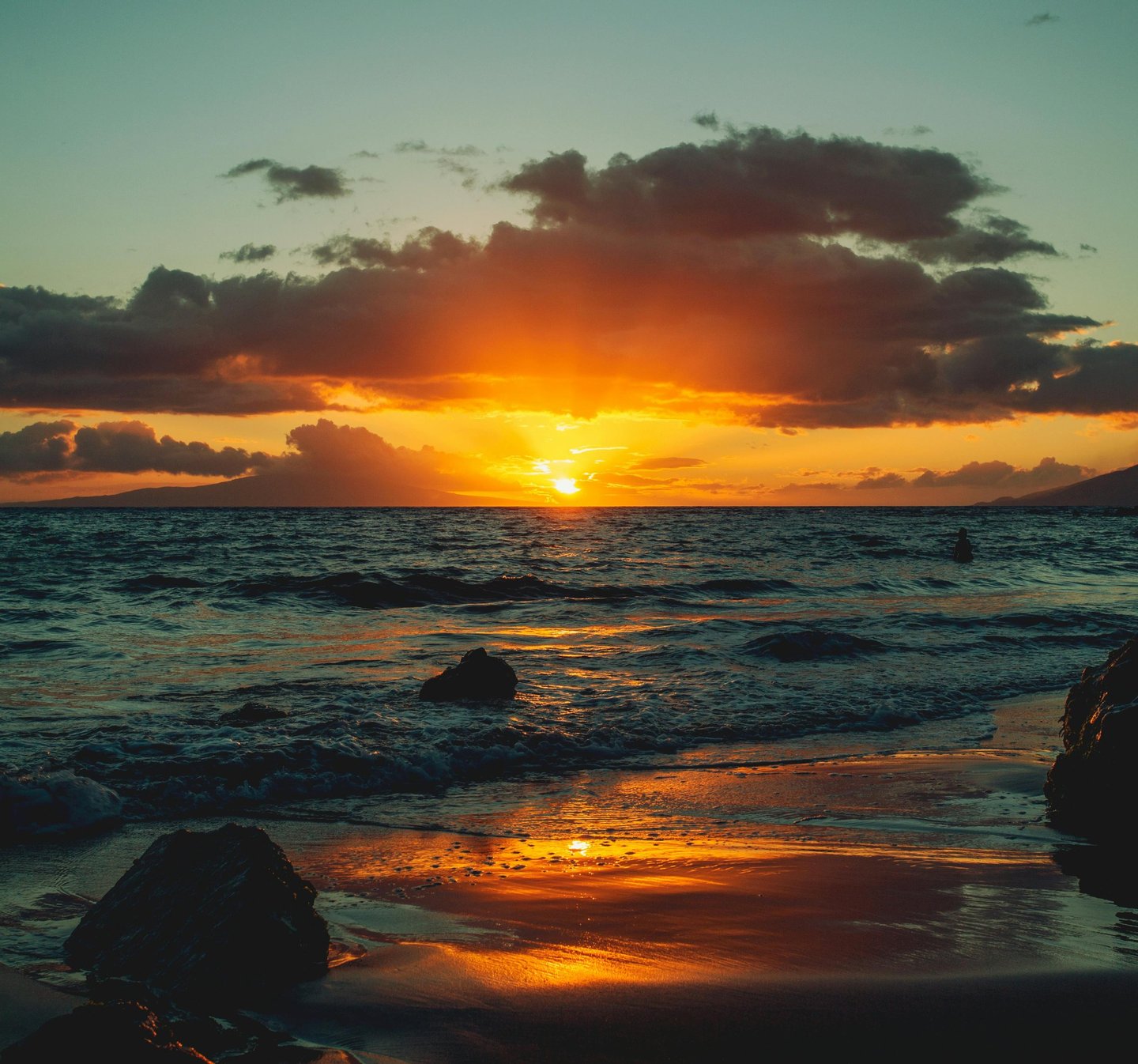 Golden tropical sunset over a dark sandy beach with ocean waves and dramatic clouds.