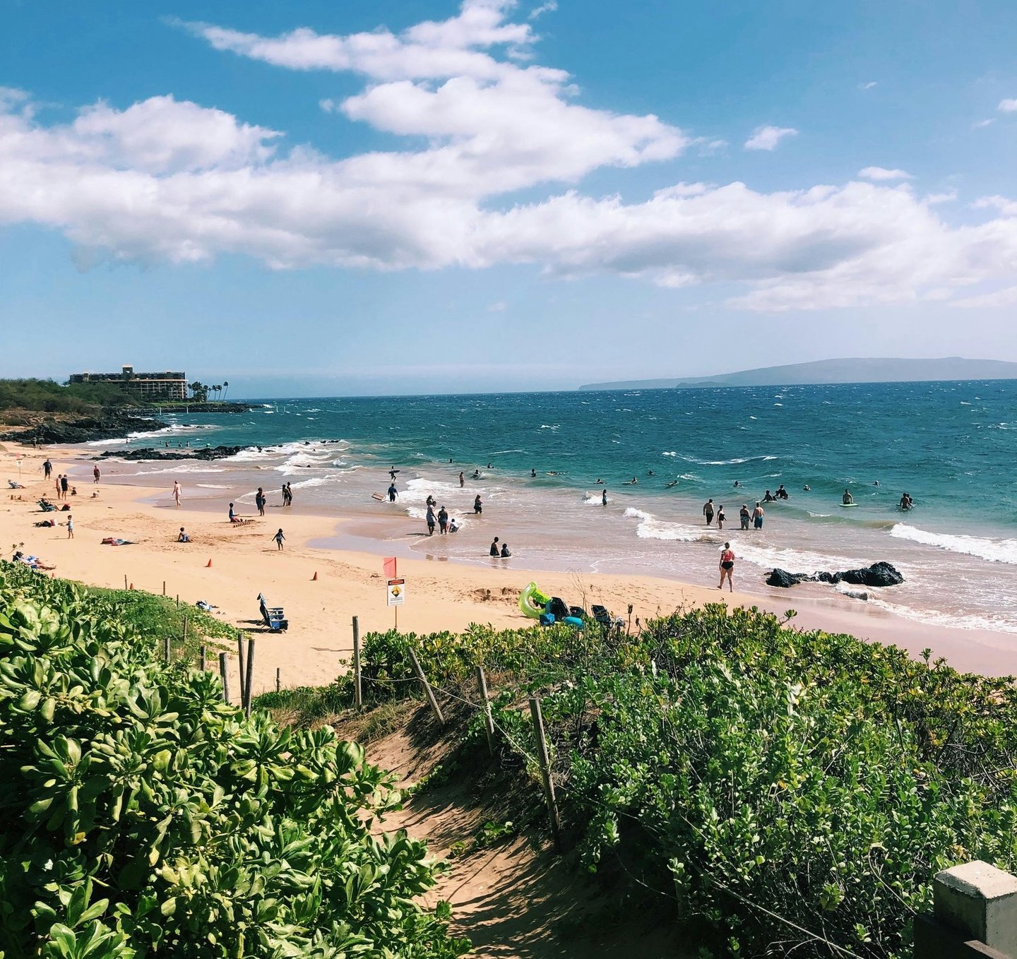 Crowded tropical beach in Maui with people swimming in turquoise ocean water under a blue sky.