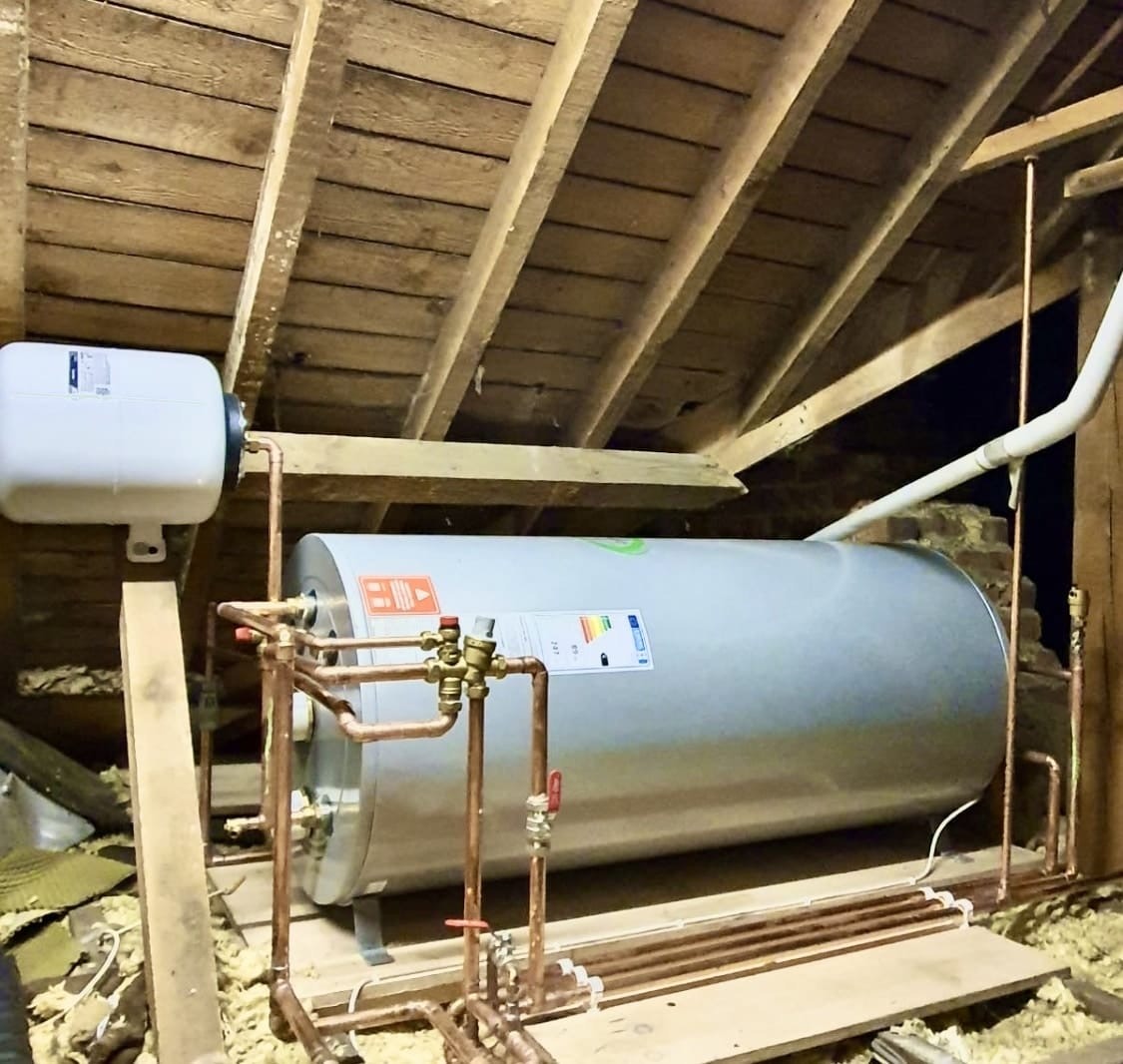 A horizontal unvented hot water cylinder installed in a wooden attic with copper pipework and expansion vessel.