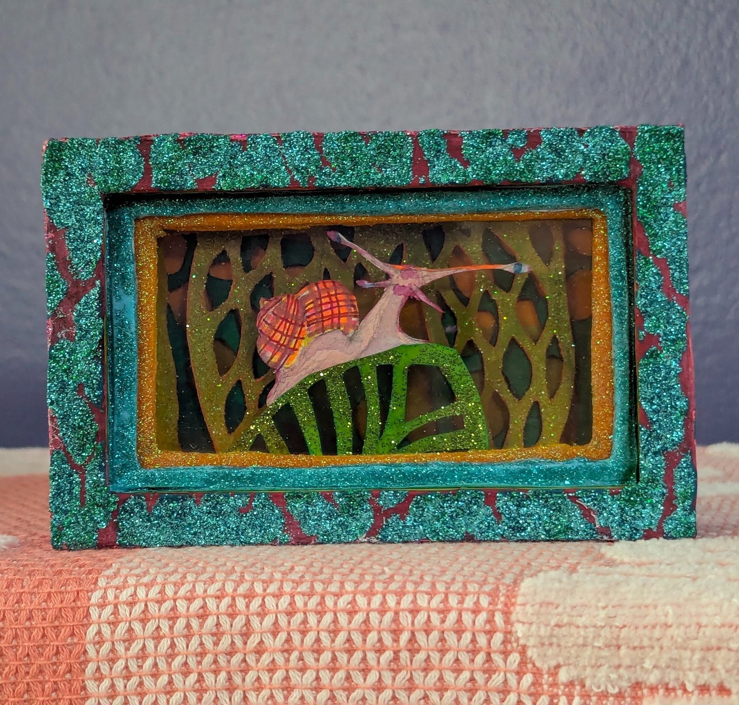 Diorama box featuring a colorful snail crawling on a leave in front of glittered layered leaf and floura affects.
