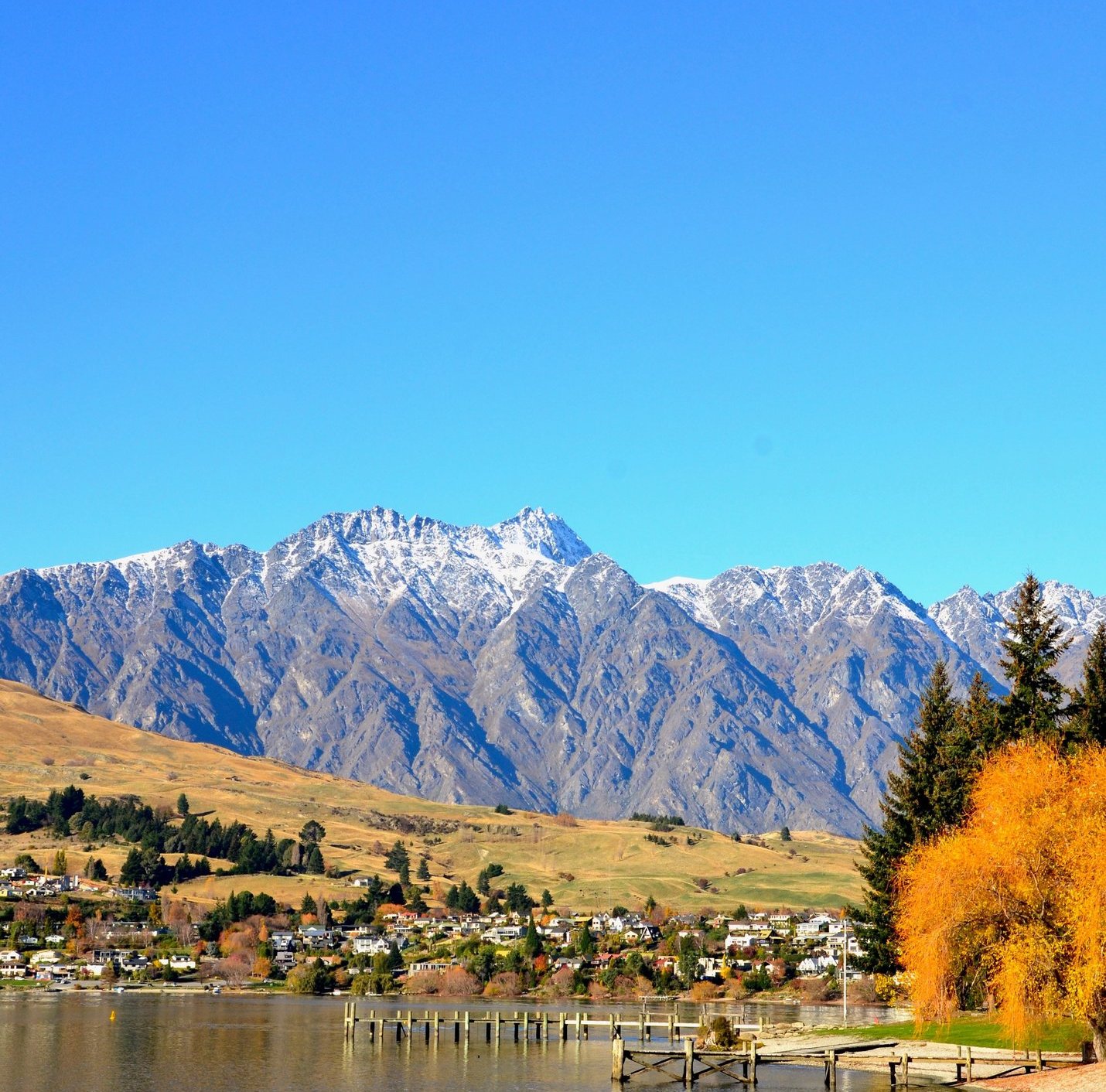 Scenic view of Lake Wakatipu with snow-capped mountains and autumn trees in Queenstown, New Zealand.
