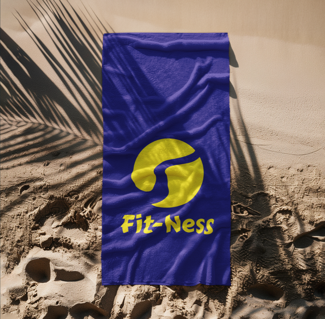 Blue Fit-Ness branded beach towel on sand under palm tree shadows.