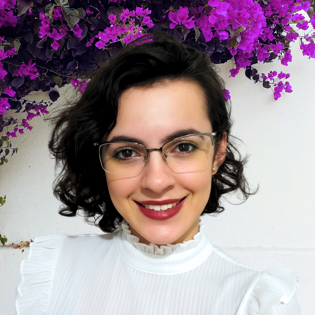 Smiling woman with short dark curls and glasses in front of purple flowers.